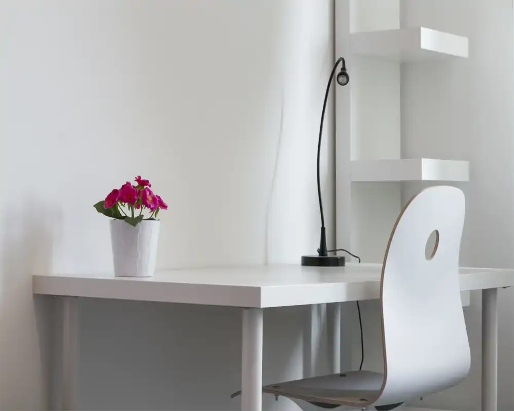 Angolo scrivania minimalista bianco con sedia moderna, lampada da lavoro e piccolo vaso fiorito che aggiunge colore — spazio di lavoro luminoso e ordinato, ideale per brevi soggiorni o lavoro da remoto.