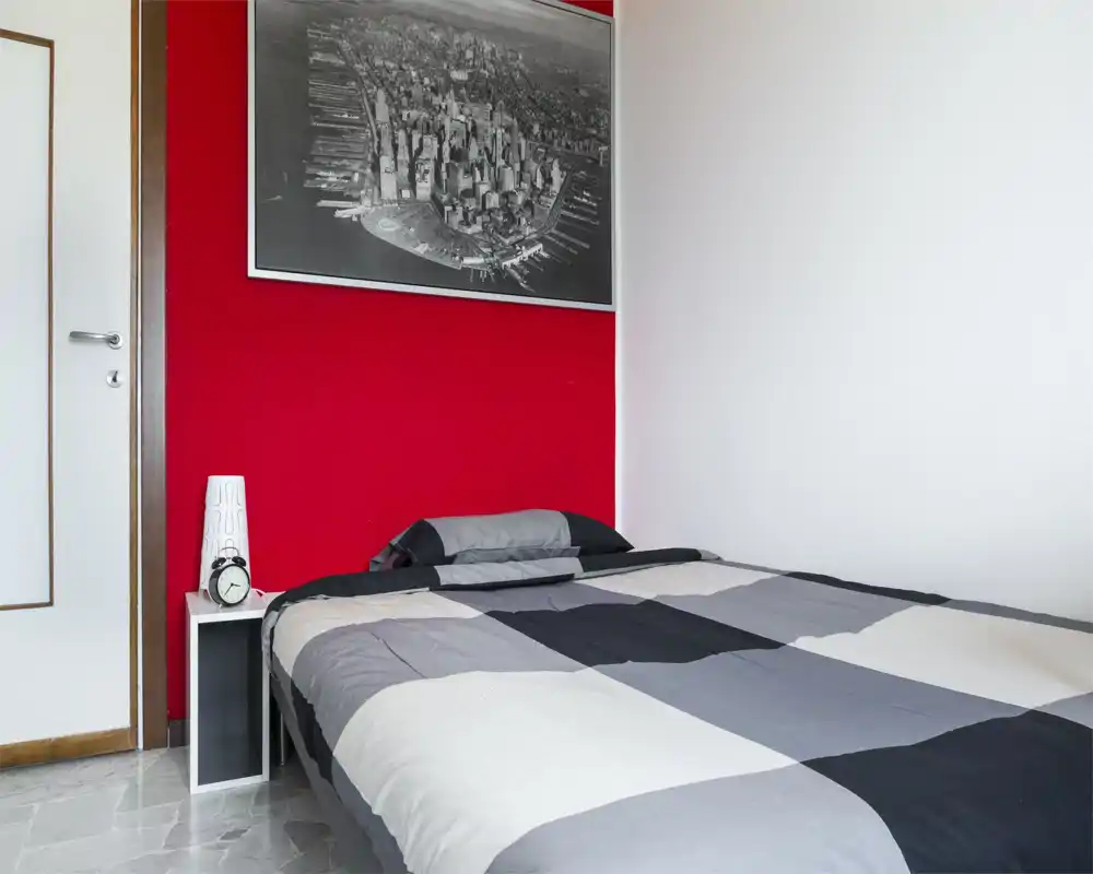 Camera da letto moderna e luminosa con letto matrimoniale con copriletto grigio a motivo geometrico, parete accentuata rossa e un piccolo comodino bianco con lampada e sveglia.