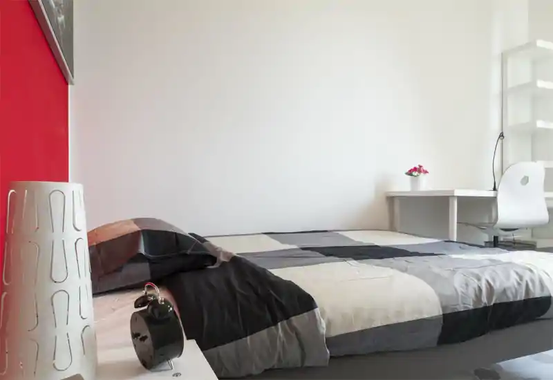 Camera da letto moderna e luminosa con un letto matrimoniale in toni grigio e nero, una lampada da comodino e sveglia, e una piccola scrivania con sedia e pianta vicino alla parete.