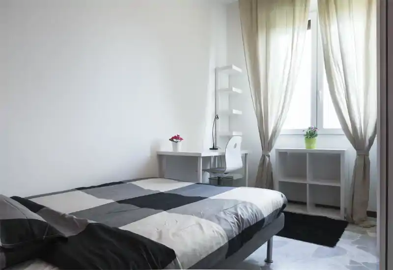 Camera da letto luminosa e minimal con letto matrimoniale in biancheria grigia, una scrivania e una sedia accanto alla finestra con tende leggere e piante in vaso.