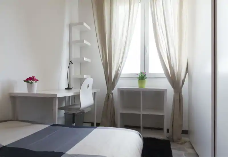Camera da letto luminosa e minimale con letto parzialmente visibile in primo piano, scrivania e sedia ordinate accanto a una grande finestra con tende leggere e piccoli vasi di fiori.