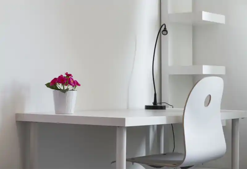 Coin bureau minimaliste blanc avec chaise moderne, lampe de travail et petit pot de fleurs apportant une touche de couleur — espace de travail lumineux et rangé, idéal pour courts séjours ou télétravail.