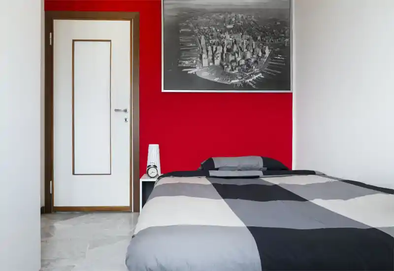 Camera da letto minimalista con un grande letto in primo piano, parete in rosso acceso, quadro della città e piccolo comodino con lampada e sveglia. Stile pulito e luminoso, adatto per annunci di affitto.