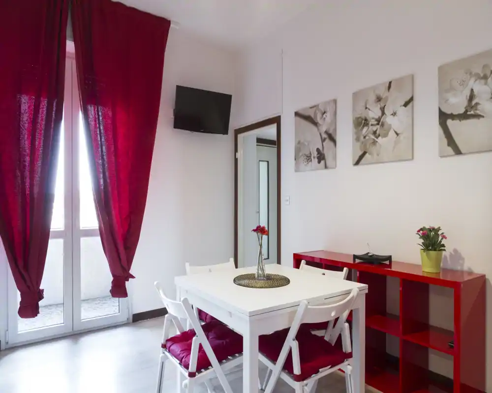 Zona pranzo compatta e luminosa con tavolo bianco e sedute con cuscini rossi, vaso di fiori decorativo e scaffale rosso abbinato; ampia finestra con tende rosse che regala luce naturale.