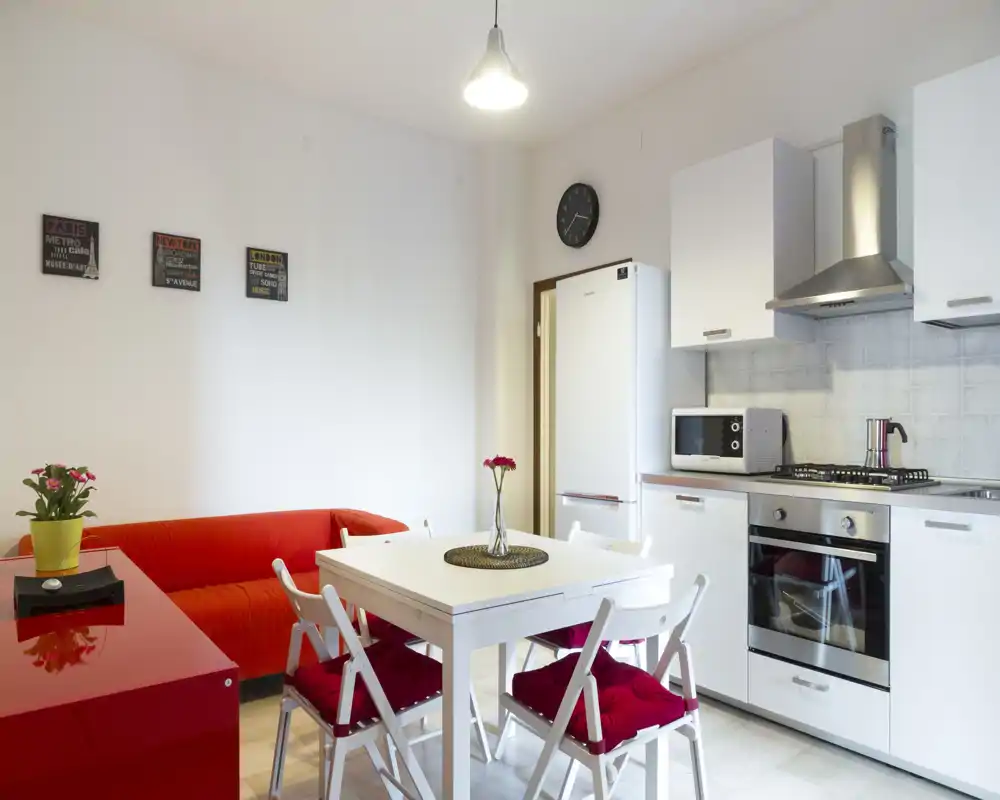 Cucina open space luminosa e moderna con pensili bianchi, forno incassato, frigorifero e un piccolo tavolo da pranzo con quattro sedie; il divano rosso e i dettagli colorati donano vivacità.