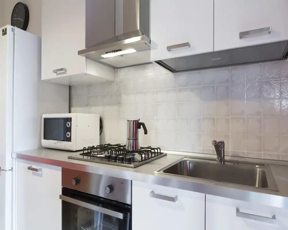 Cucina moderna e compatta con piano in acciaio, piano cottura a gas, forno, microonde e lavello singolo — luminosa, pulita e ordinata.