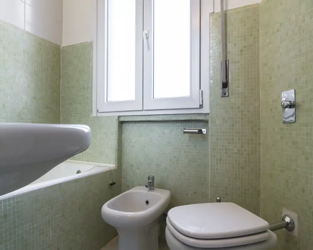 Bagno compatto con piastrelle a mosaico verdi, bidet, WC e vasca sotto la finestra; luce naturale intensa e sanitari puliti.