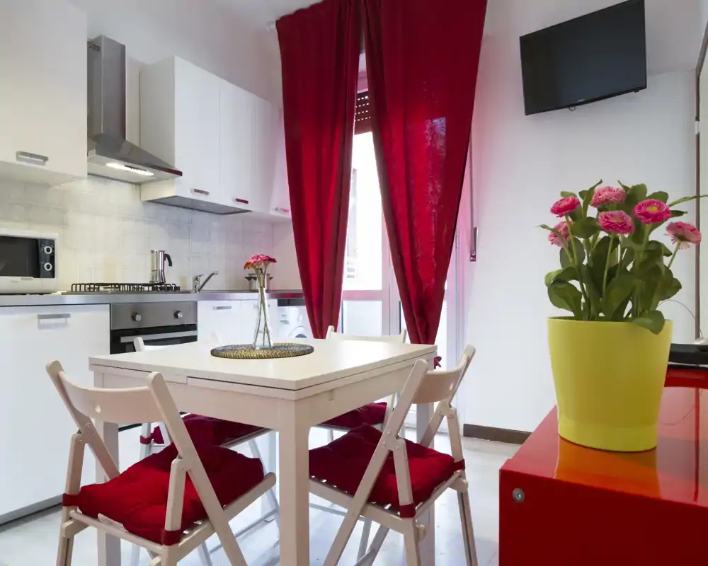 Cucina moderna e luminosa con un piccolo tavolo da pranzo e quattro sedie con cuscini rossi e tende coordinate; perfetta per pasti accoglienti in un appartamento compatto.
