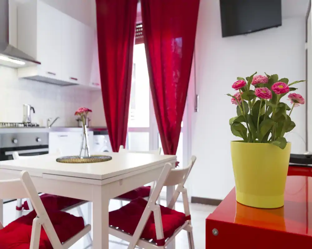 Cucina luminosa con tavolo da pranzo bianco e accenti rossi; elettrodomestici moderni, cuscini accoglienti e fiori decorativi rendono l'ambiente invitante.
