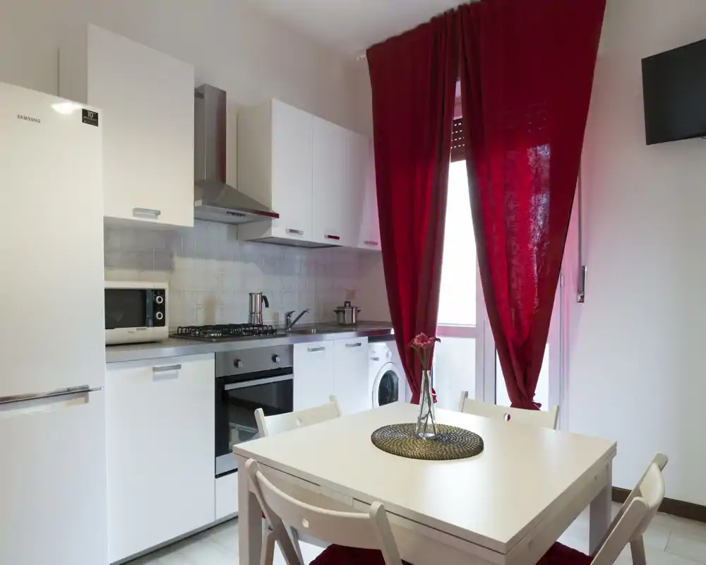 Cucina moderna e luminosa con pensili bianchi, forno incassato, piano cottura a gas e un piccolo tavolo da pranzo. Le tende rosse e il centrotavola semplice creano un’atmosfera accogliente, ideale per affitti brevi.