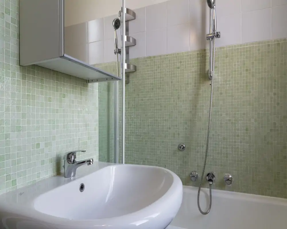 Bagno compatto e pulito con lavabo bianco, rubinetto cromato e doccia sopra la vasca; piastrelle mosaico verde chiaro donano un tocco fresco.