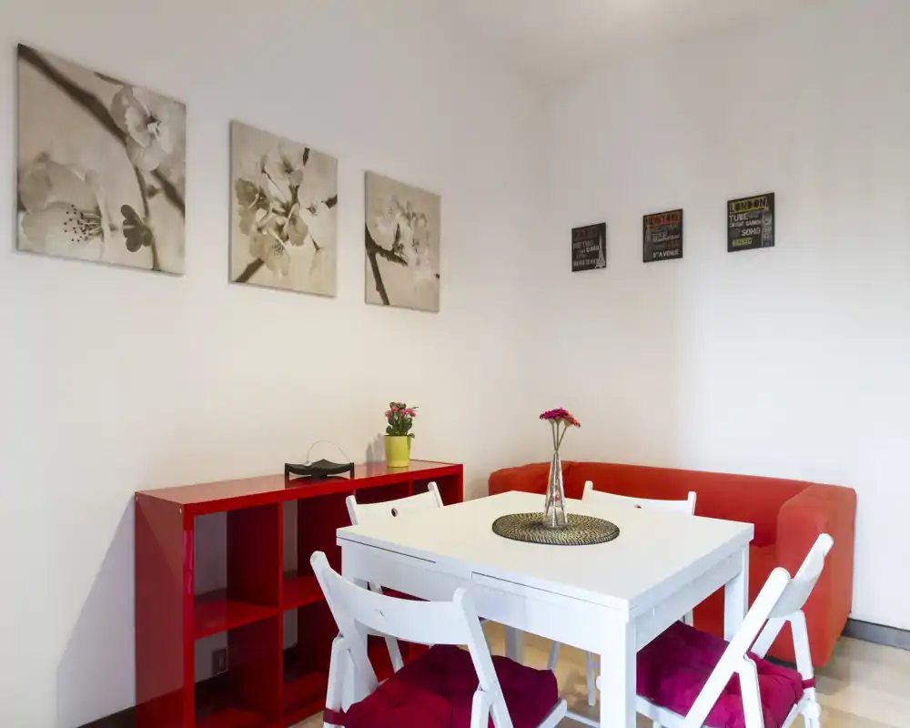 Angolo living/pranzo luminoso e curato con tavolo bianco, divano rosso e quadri decorativi — perfetto per un monolocale compatto.