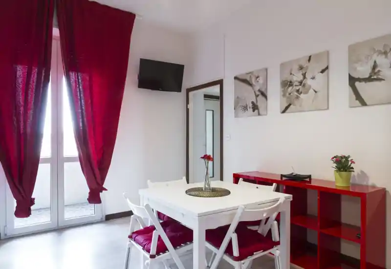 Coin repas lumineux et compact avec table blanche, chaises aux coussins rouges, fleurs décoratives et étagère rouge assortie ; grande fenêtre avec rideaux rouges apportant une forte lumière naturelle.