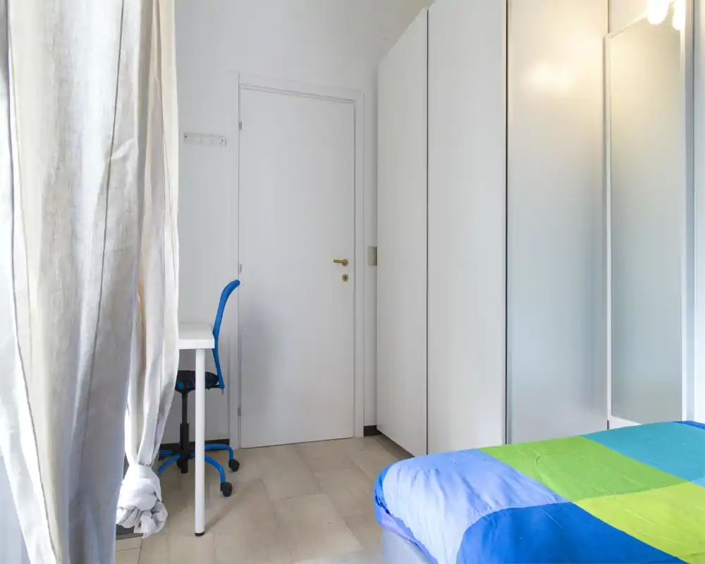 Bright, minimal bedroom with a colorful blue-green bedspread, white wardrobe and a small desk with a blue chair near the door.