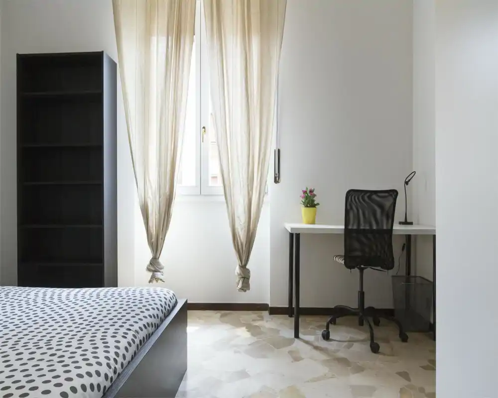 Minimalist bedroom with a polka-dot bedspread, a shelving unit, and a bright workspace by the window — airy natural light and clean lines ideal for rental photos.