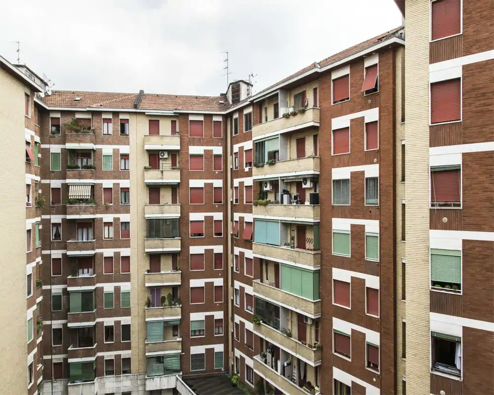 Exterior view of a residential building courtyard showing multiple apartment facades, balconies and closed shutters. The photo captures the building exterior rather than any specific indoor room.