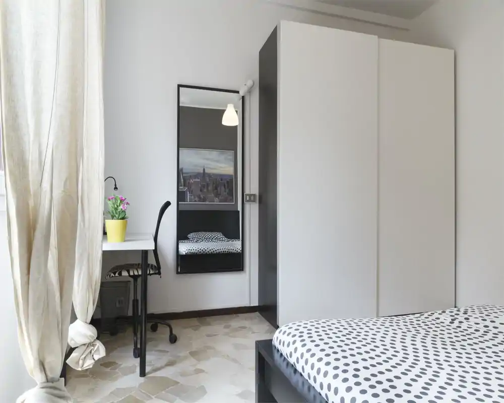Bright, minimalist bedroom with a polka-dot bedspread, large wardrobe and a desk by the window. The room is well-lit with natural light and simple, modern furnishings ideal for a rental listing.