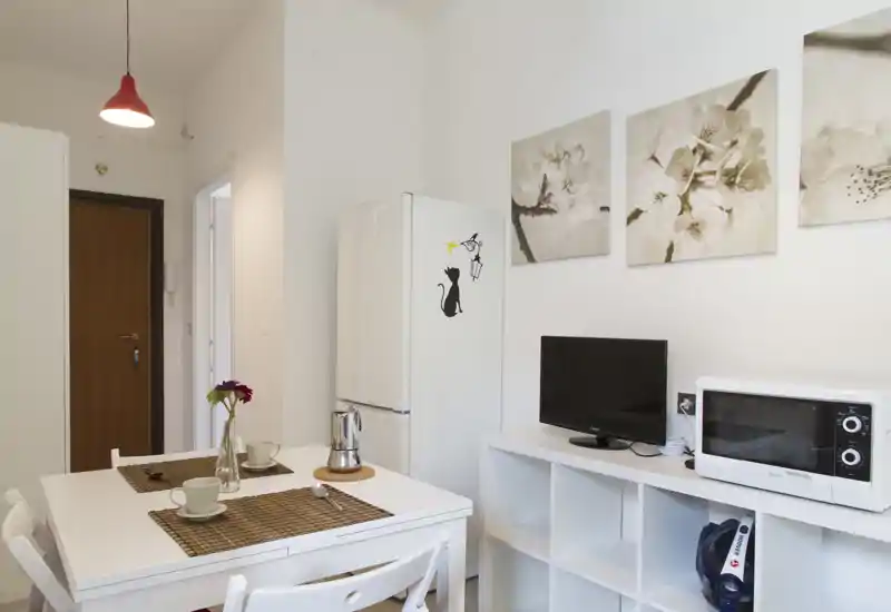 Compact open kitchen with a white dining table set for two, refrigerator, microwave and small TV; clean, bright and simply decorated for a cozy stay.