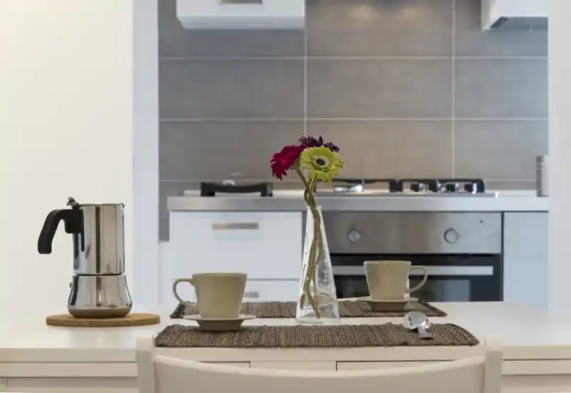Bright, modern kitchen corner with a small dining surface set for two: two cups, a moka pot and a vase with flowers in front of a built-in oven and stovetop.