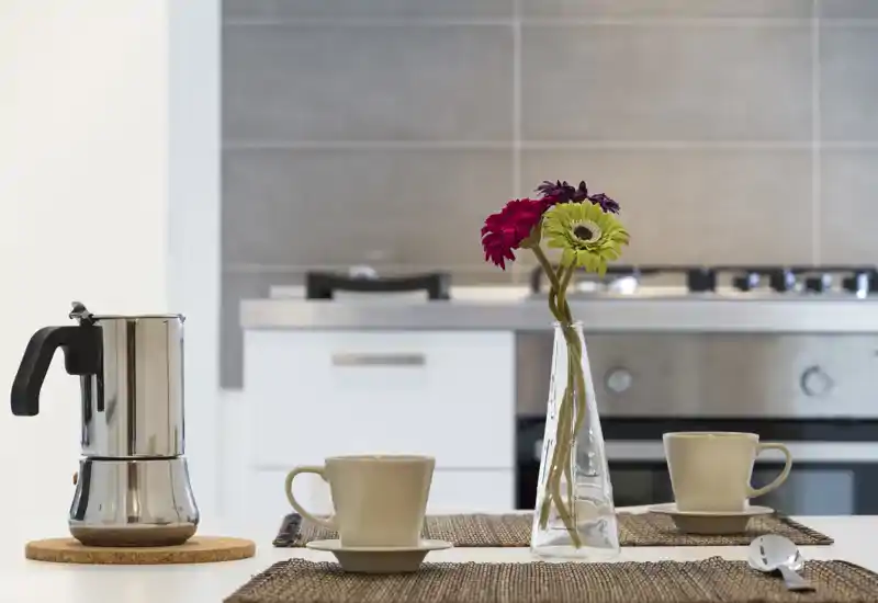 Bright, modern kitchen scene with a stovetop and oven in the background and a simple table setting in the foreground featuring cups, a moka pot and a vase with fresh flowers.