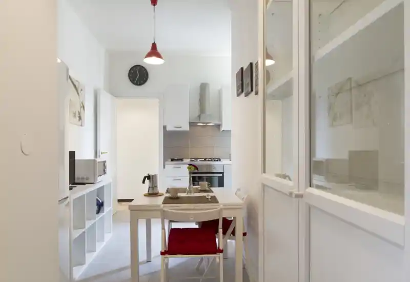 Compact modern kitchen with a small dining table set for two, white cabinetry, stainless-steel range hood and warm decorative touches that create a bright, inviting space.