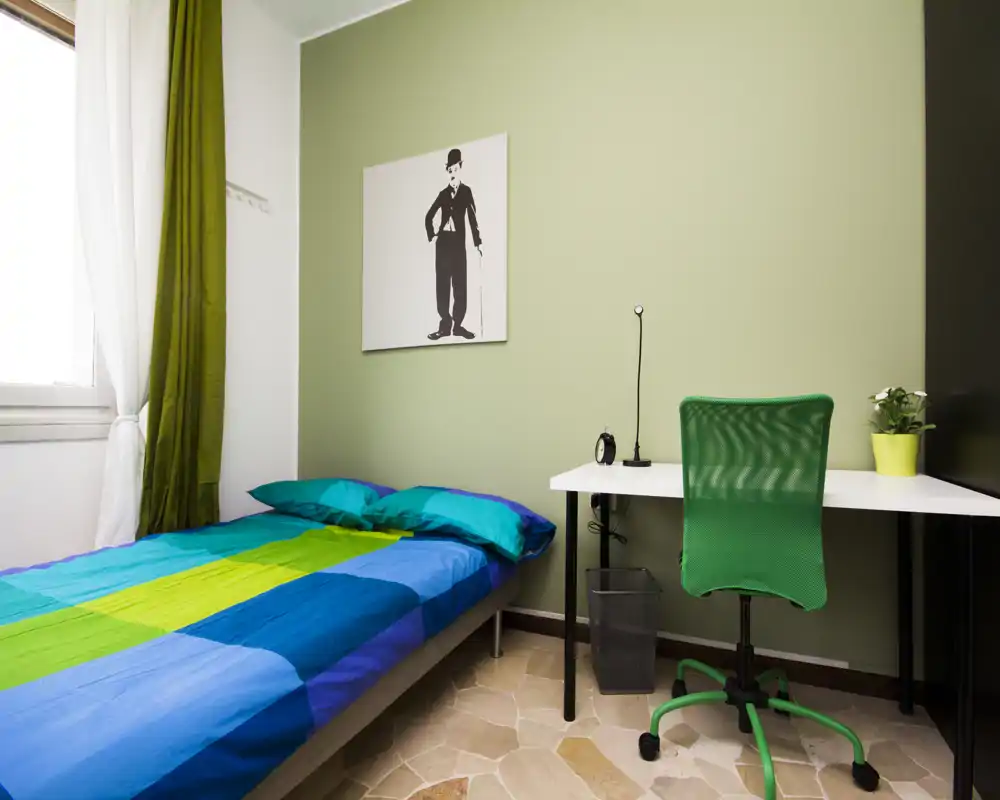 Bright, neatly made single bedroom with colorful bedding, a desk and ergonomic chair — ideal for a student or single occupant.