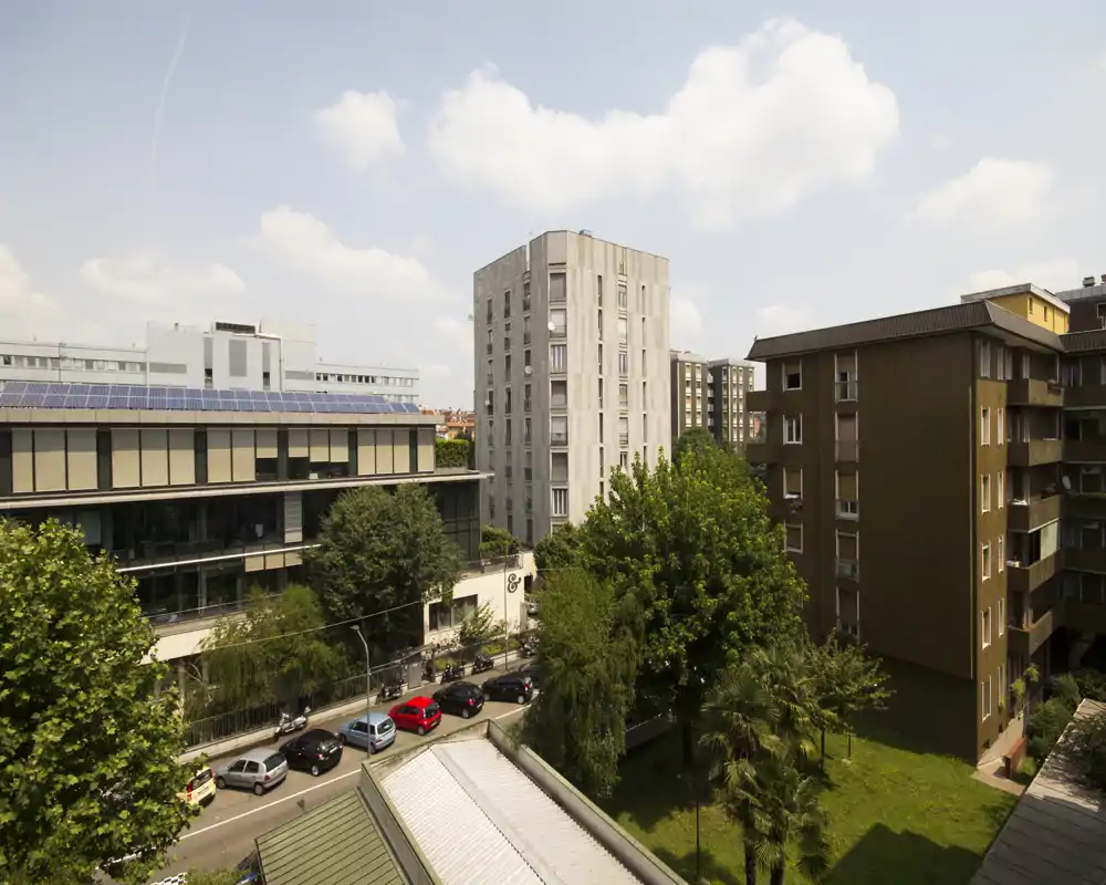 Sunny urban view from a balcony or window showing neighboring apartment buildings, trees and a street with parked cars under a blue sky.