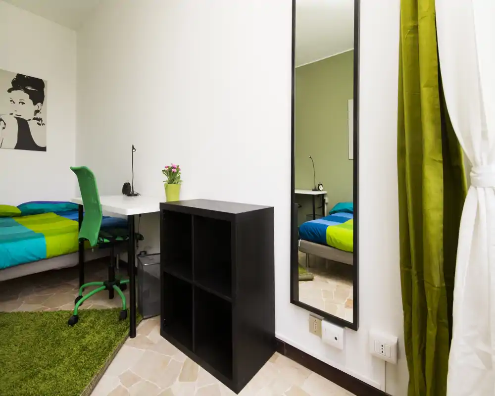 Bright, neatly staged bedroom corner with a colorful bed, desk and mirror — ideal for single occupancy. The room features a green-accented chair and rug, a small desk with lamp, and a full-length mirror that enlarges the sense of space.