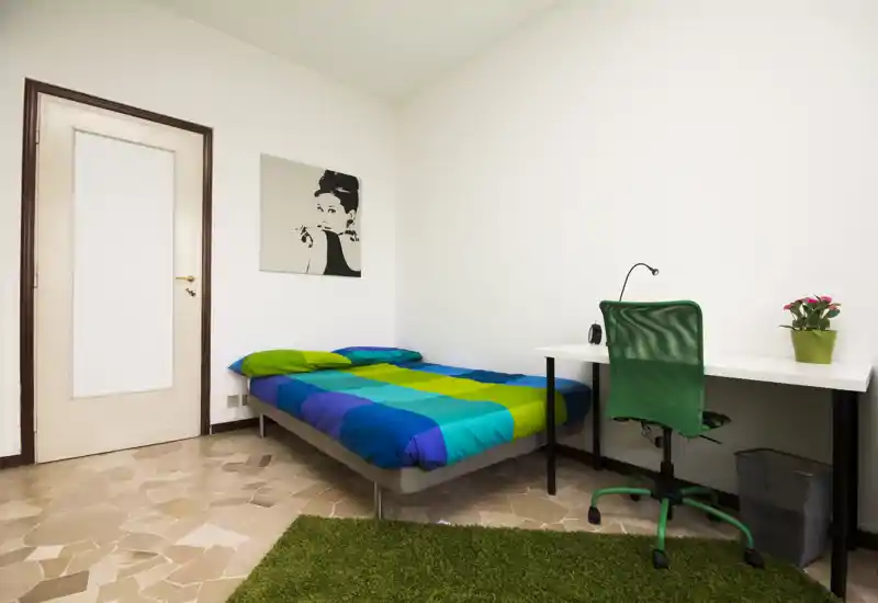 Bright, modern bedroom with a colorful double bed, a simple white desk and ergonomic green chair beside a small plant; neutral walls and stone-tiled floor create a clean, minimalist feel.