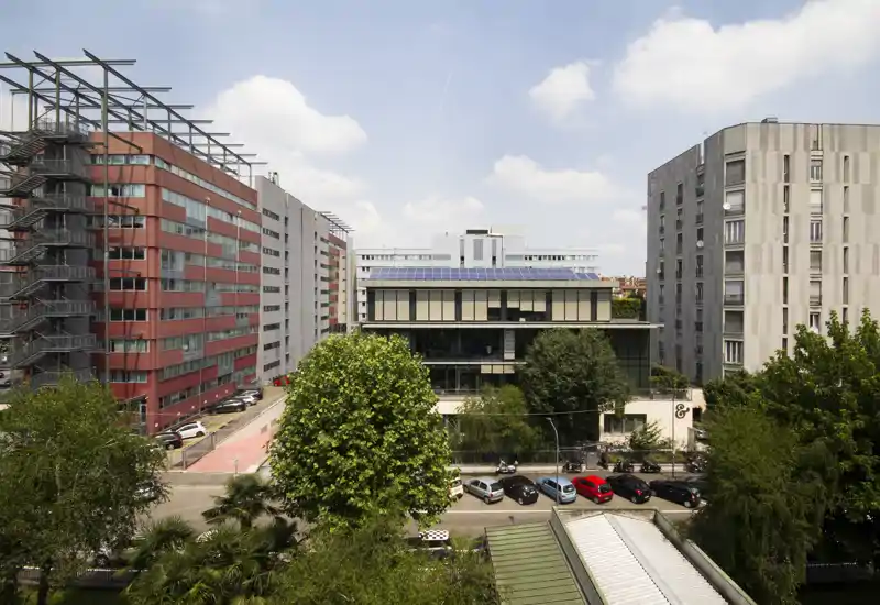 City exterior view with modern buildings, trees and parked cars under a bright blue sky — suitable as an exterior/urban listing photo.
