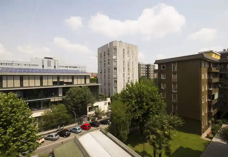Sunny urban view from a balcony or window showing neighboring apartment buildings, trees and a street with parked cars under a blue sky.