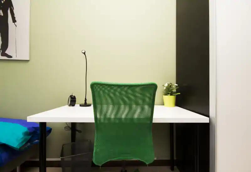 Small bedroom corner with a white desk and a bright green mesh chair facing the camera; a portion of the bed with blue bedding is visible at left and a small potted plant and lamp sit on the desk.