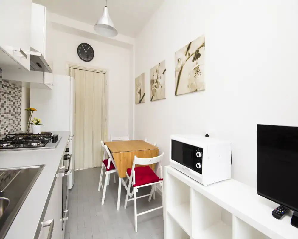 Small modern kitchen with a gas hob, sink and compact dining table for four. Bright, neutral decor with wall art and red seat cushions adding a friendly touch.