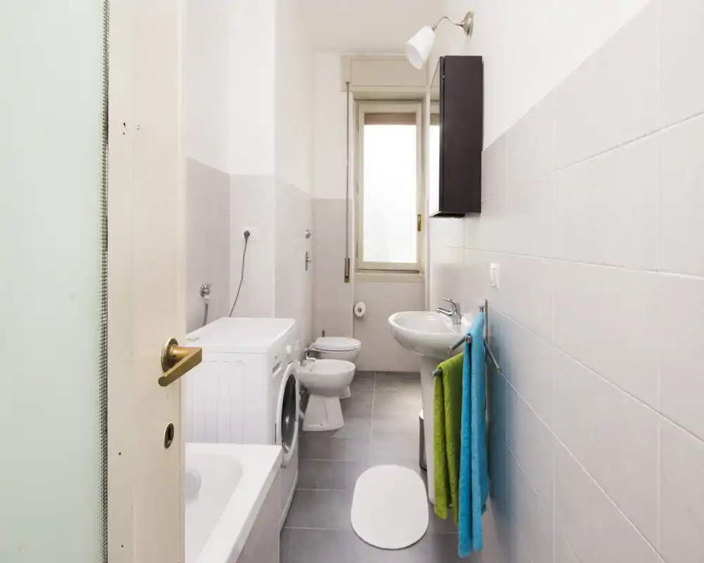 Bright, modern bathroom with a bathtub, washing machine, bidet, toilet and sink; light tiles and a window provide natural light.