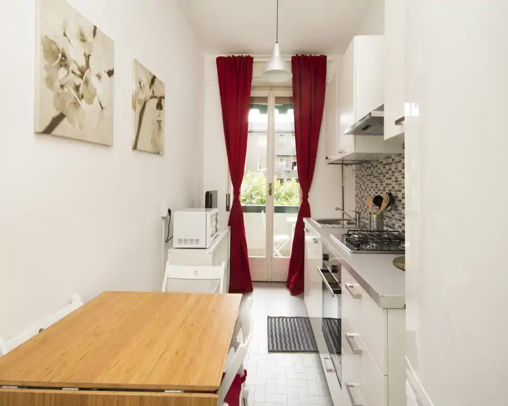 Bright and compact modern kitchen with white cabinets, gas hob, and a wooden dining table next to a balcony door with red curtains.