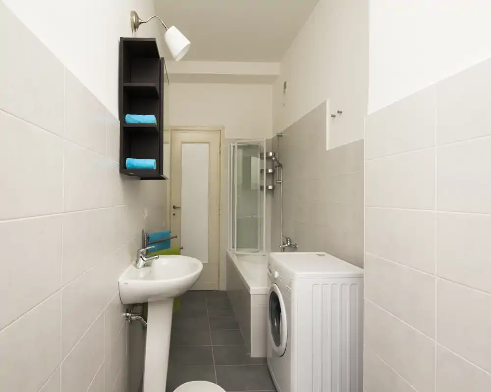 Clean, narrow bathroom with a pedestal sink, bathtub with glass shower screen and a washing machine; neutral tiles and bright lighting create a neat, functional space.