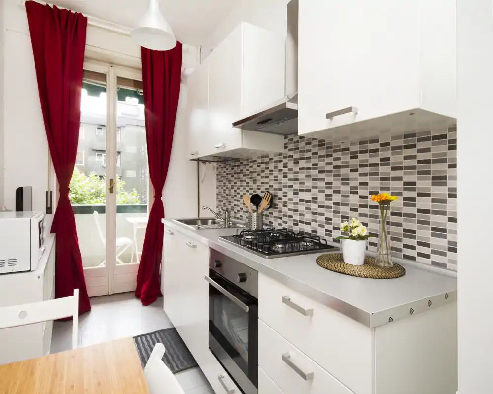 Modern, compact kitchen with white cabinets, stainless countertop, gas hob and oven. Natural light streams through glass doors opening onto a small balcony; red curtains add a warm accent.