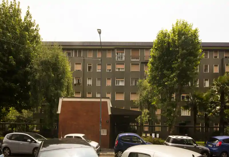 Exterior view of an apartment block with parked cars, trees and a small brick entrance building; suitable for showing the building façade and outdoor parking.