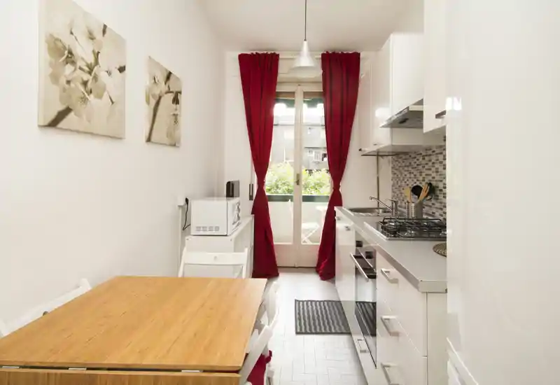 Bright and compact modern kitchen with white cabinets, gas hob, and a wooden dining table next to a balcony door with red curtains.