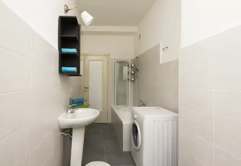 Clean, narrow bathroom with a pedestal sink, bathtub with glass shower screen and a washing machine; neutral tiles and bright lighting create a neat, functional space.