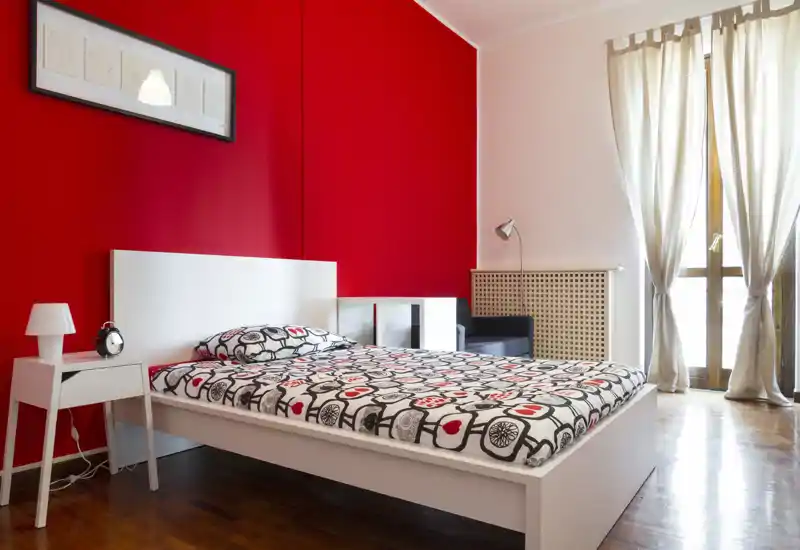 Dormitorio moderno y luminoso con cama blanca sobre plataforma frente a una pared roja intensa, ropa de cama estampada, mesita y gran ventana con cortinas translúcidas que dejan entrar la luz natural.