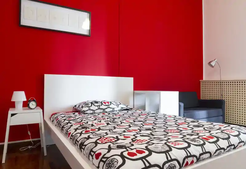 Dormitorio moderno y luminoso con cama blanca y edredón estampado frente a una pared roja intensa. La imagen destaca la cama, la mesita con lámpara y un pequeño sofá, creando un espacio atractivo y acogedor.