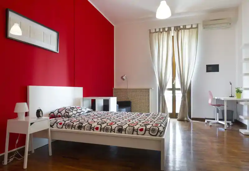 Dormitorio luminoso y moderno con pared roja, cama doble con ropa de cama estampada, mesita y un pequeño escritorio junto a la ventana — ideal para estancias cómodas.