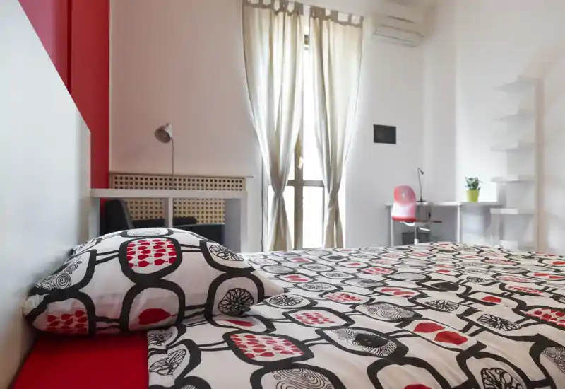 Dormitorio moderno y luminoso con ropa de cama estampada, un escritorio y luz natural de una ventana alta. Decoración limpia y un práctico rincón de trabajo.