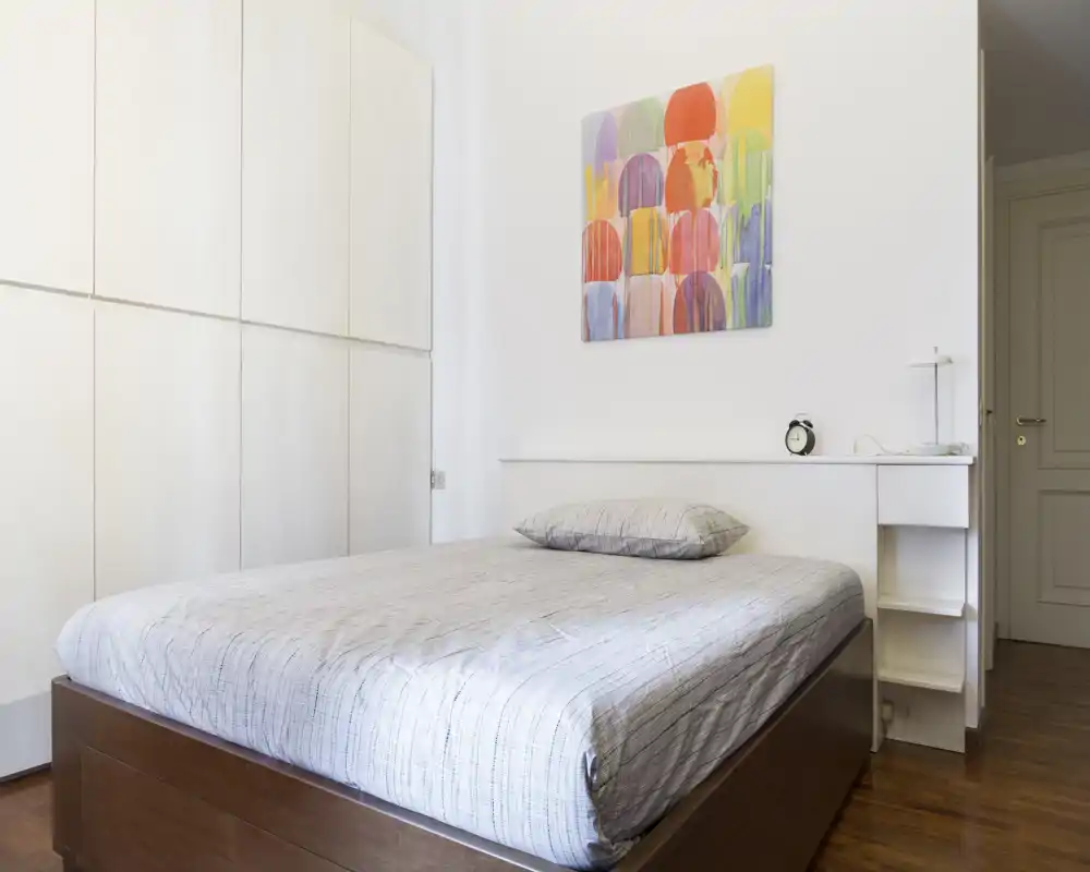 Camera da letto luminosa e moderna con ampio letto in legno, semplice comodino a mensola e quadri colorati che danno un aspetto fresco e minimalista.