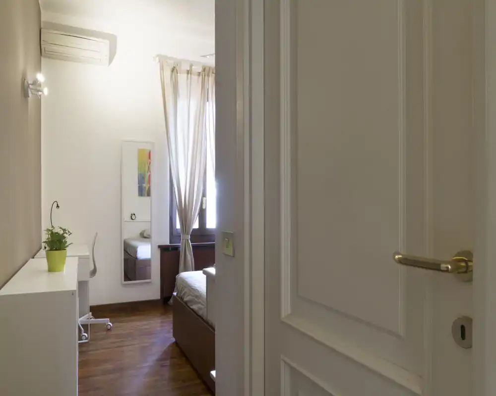 Camera da letto luminosa e ordinata vista da una porta aperta, con pavimento in legno, letto rifatto e una scrivania a muro — ideale per affitti singoli.