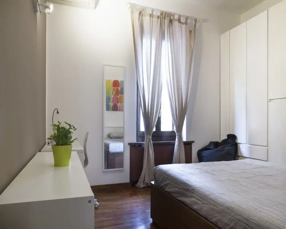 Camera da letto luminosa e moderna con letto matrimoniale visibile, ampia finestra con tende leggere, armadio a muro e una piccola scrivania — spazio confortevole e ben illuminato ideale per un annuncio.