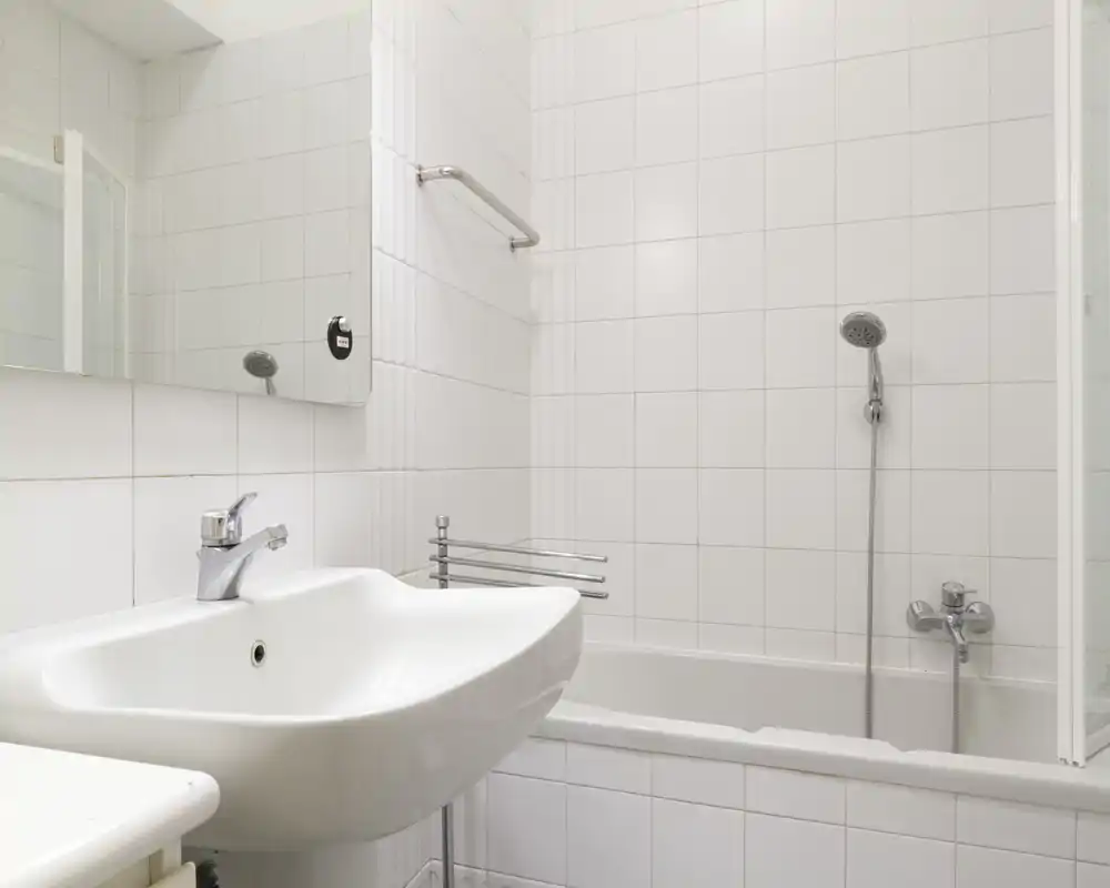 Bagno bianco e pulito con lavabo, specchio e vasca con doccia a mano; pareti piastrellate e rubinetteria cromata offrono uno spazio semplice e funzionale.