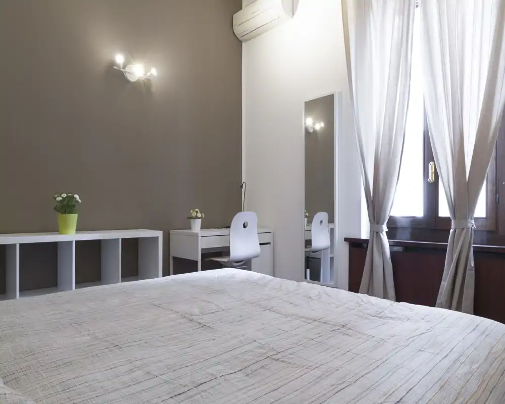 Camera da letto luminosa e semplice con un grande letto in primo piano, una scrivania e uno specchio vicino alla finestra e tonalità neutre per un'atmosfera rilassante.
