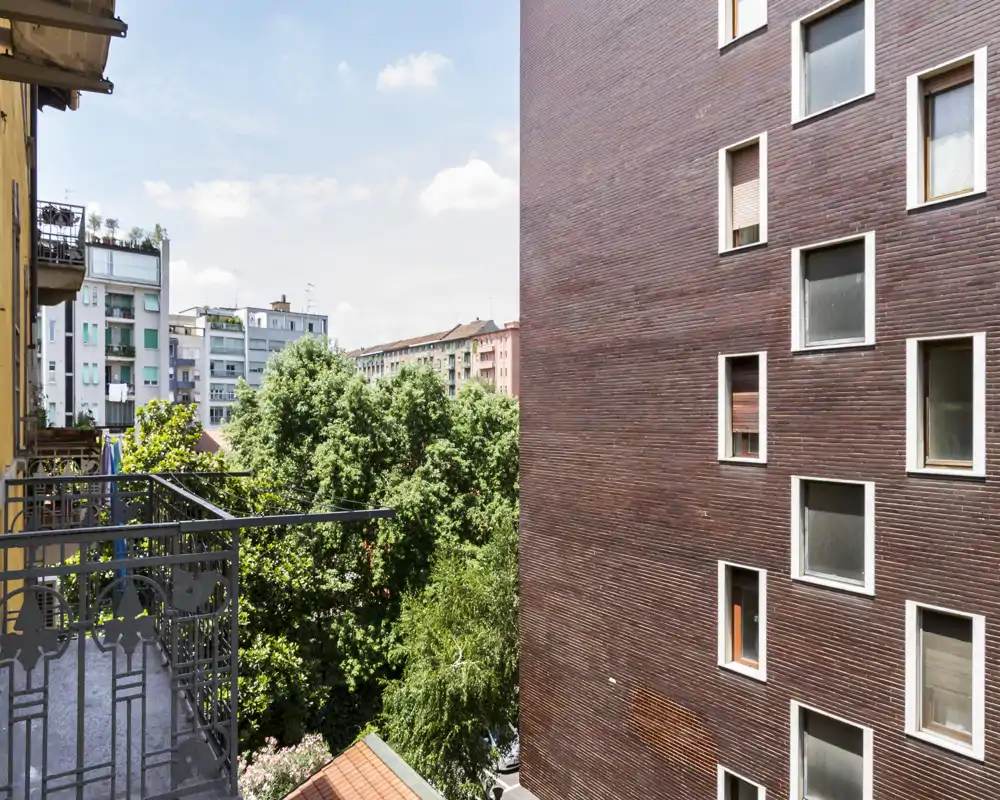 Vista dal balcone soleggiata su alberi frondosi e palazzi vicini; ringhiera in ferro in primo piano e cielo sereno.